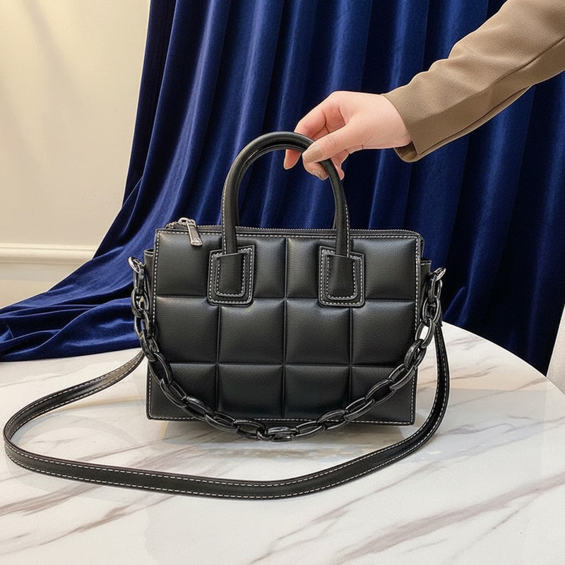 Bolso acolchado negro vestismuse con costuras blancas y cadena decorativa sobre mesa de mármol